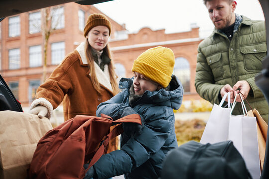 Family of three packing bags into car trunk on chilly day with snowy ground visible around, parents assisting their young child while wearing warm clothing