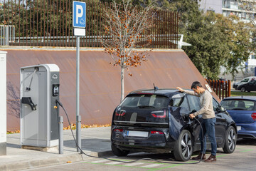 A man plugs in a dark blue electric car at a public EV charging station. Urban parking area with signage, trees, and parked vehicles in the background. Horizontal wide-angle shot with copy space.