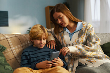 Mother explaining smartphone features to her child sitting on couch in cozy living room setting,...