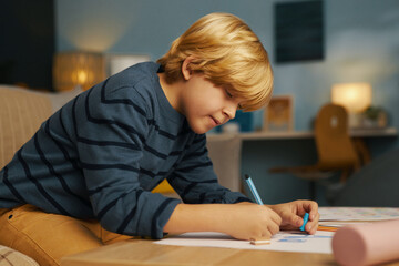 Fototapeta premium Young boy attentively drawing with colored pencils at home in warmly lit, comfortable living room, seated at table with soft background blur creating peaceful atmosphere