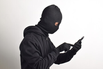 Mysterious man wearing black hoodie and mask doing hacking activity on mobile phone, hacker holding...