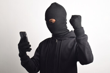 Mysterious man wearing black hoodie and mask doing hacking activity on smartphone, with successful...