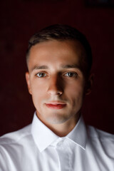 a man with short dark hair and fair skin. He has brown eyes and neat facial features. He is wearing a white shirt and looking straight into the camera. 