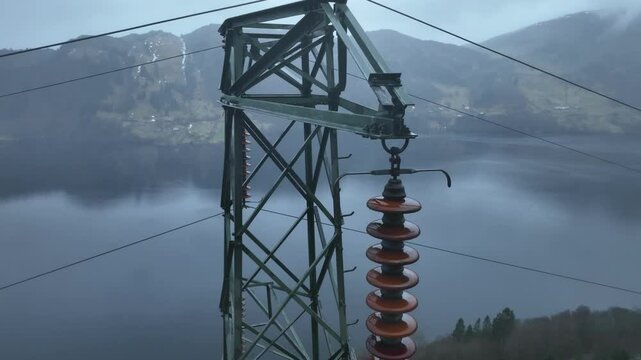 High voltage pylon top with insulators in focus. Drone descends slowly while tilting up, creating parallax against a fjord background in rainy, misty weather