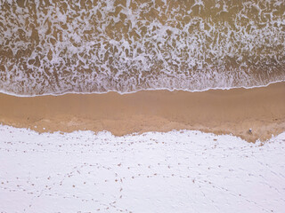 An overhead perspective of a winter shoreline where foamy ocean waves meet snowy land. The combination of textures and colors highlights the beauty of seasonal transitions in nature.