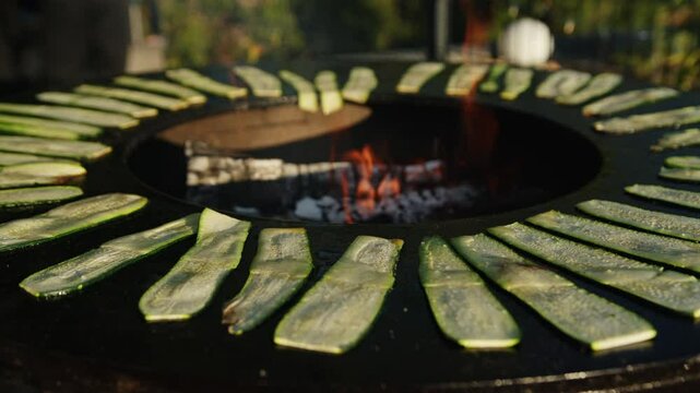 Slices of fresh zucchini are arranged in a circular fashion on a hot grill, sizzling gently as they cook over glowing embers. The outdoor environment is filled with greenery and warmth.