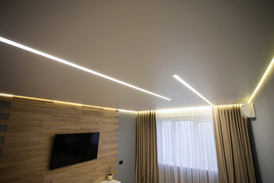 Beautiful modern stylish ceiling with light lines against the background of a window with curtains and a TV in the living room. Stylish interior.