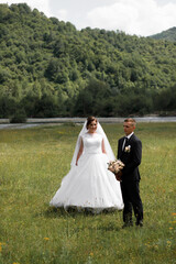 Newlyweds against the backdrop of nature. The groom is in a black suit and white shirt, the bride is in a white dress with lace and a veil.