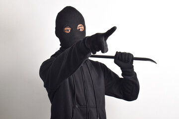 Thief wearing black jacket and mask holding crowbar, posing pointing at camera, isolated on white...