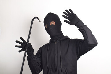 Thief wearing black jacket and mask holding crowbar, surrendering with hands up, isolated on white...