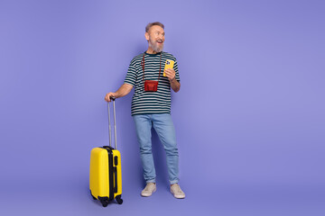 Happy elderly man with a yellow suitcase and a smartphone standing against a purple background,...