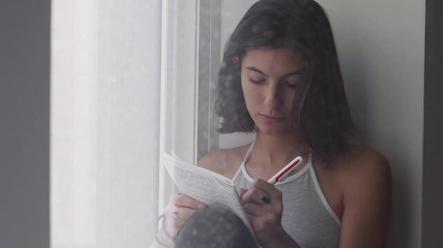A young woman with black hair carefully writes an important letter by the window, feeling passionate and emotional. She hugs the letter at the end.