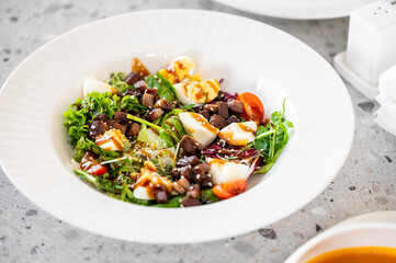 Fresh mixed salad with greens, cherry tomatoes, boiled eggs, and diced meat on a white plate, served on a speckled surface, showcasing a healthy and appetizing meal.