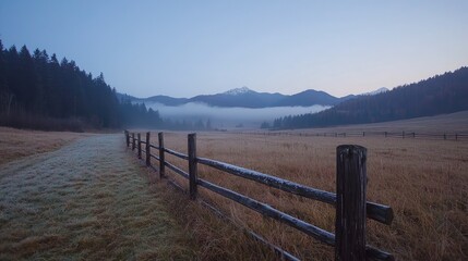 Obraz premium Wooden fence runs along a frosty dirt path in a misty mountain valley during a quiet autumn dawn.