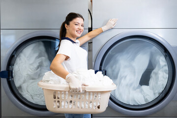 Smiling female worker holding laundry basket by washing machine in dry cleaning service.