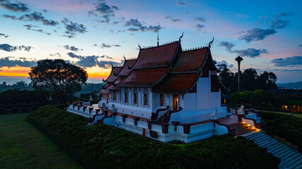Obraz premium Aerial view of a traditional temple surrounded by greenery under a vibrant sunrise sky. The golden and blue hues of twilight highlight the intricate architecture and peaceful ambiance of the scene