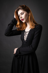 Red-haired gothic girl in a black dress on a gloomy background in the studio.