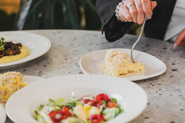 A close-up of a person's hand holding a fork to cut a creamy dish on a white plate, with colorful salads in the foreground and a stylish dining table setting.