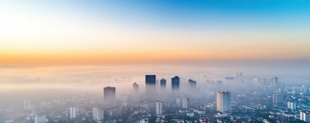 Obraz premium A panoramic cityscape shrouded in morning fog, with skyscrapers peeking through beneath a vibrant sky transitioning from blue to warm hues.