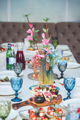 A table with a vase of flowers in the center. The table is set for a meal with a variety of dishes and drinks