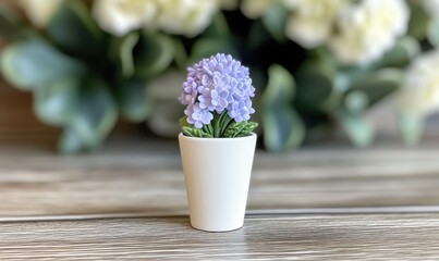 A small white pot with purple flowers placed on a wooden surface, surrounded by blurred greenery