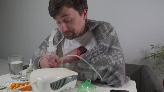 A young man sits at home wrapped in a blanket, suffering from a sore throat and cough. He uses a nebulizer, a medical device for inhalation therapy, to treat cold, bronchitis, and asthma symptoms