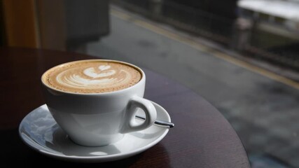 A steaming cup of coffee rests on a table with intricate latte art. Outside, cars pass by, capturing the energy of a lively urban setting, showcasing daily life in motion.