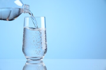 Pouring refreshing soda water from bottle into glass on light blue background, closeup. Space for text