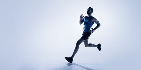 Dynamic image of muscular young man, runner in blue jersey and black shorts, training, preparing for marathon. Concept of sport, competition, endurance, athletics, sportive lifestyle. Copy space