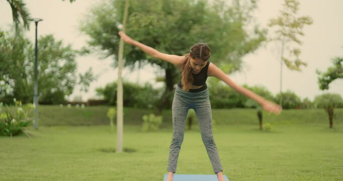 Indian young adult gen z sporty woman lady do self health care warm up on mat in fresh air park garden sports teen girl female precise cross body bend toe touch yoga asana learn day time good habit