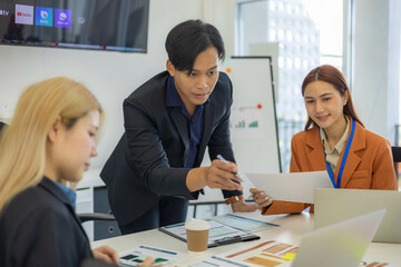 Three people are sitting at a table with a laptop open. One of them is laughing while the other two look on. The man in the middle is giving a presentation to the other two
