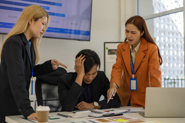 Three people are in a meeting room, with one of them pointing at a laptop screen. The man on the laptop is looking down, and the other two people are looking at him. Scene is tense and serious
