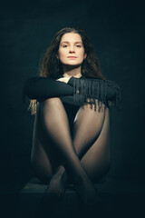 Fototapeta premium Sensuality young girl with crossed legs, curling hair in a black blouse against a black background in light, black and white dramatic portrait with light beam. 