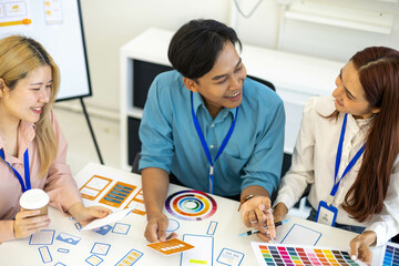 Three people are sitting around a table, working on a project. They are wearing name tags and are pointing at a whiteboard. Scene is collaborative and focused
