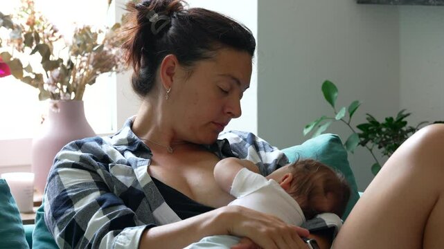 Mother breastfeeding baby on couch in living room, nurturing maternal bond, woman holding infant close in relaxed home environment, plants and flowers in background