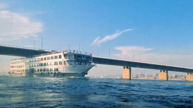 A white luxury cruise ship glides on the blue Nile River, passing under a modern bridge, Aswan to Luxor, Egypt. The clear blue sky and distant cityscape enhance the scenic journey.