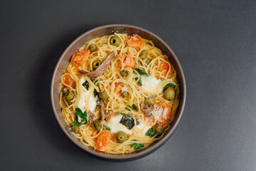 Flat lay of Spaghetti pasta in a bowl served with olives, tomatoes, cheese, and herbs, plated on a clean dark surface for an appetizing and elegant composition
