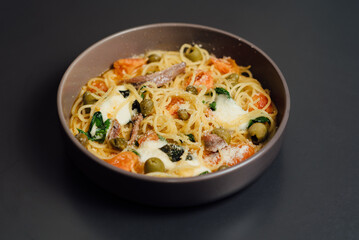 Spaghetti pasta in a bowl served with olives, tomatoes, cheese, and herbs, plated on a clean dark surface for an appetizing and elegant composition