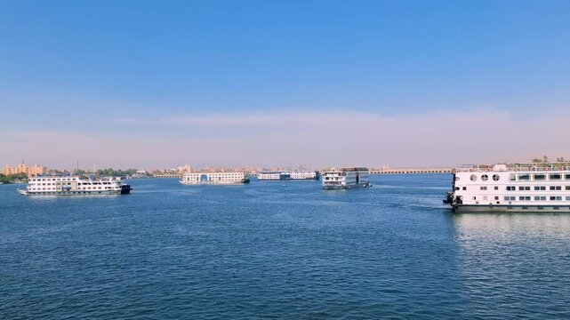 Multiple white cruise ships glide on the blue Nile River, under a clear blue sky, near Esna Lock, Egypt. The distant cityscape and calm waters create a picturesque travel scene.