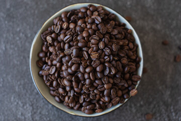 Pile of coffee beans in a bowl spilling over the side on a dark and moody background. Overhead top shot