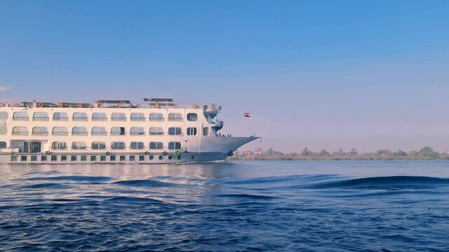 A white cruise ship with multiple decks sails on the calm blue Nile River. The Egyptian flag flying high and the historical significance of the route, Aswan to Luxor, Egypt.