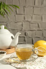Refreshing green tea in cup, lemon and dry leaves on white table against grey textured wall