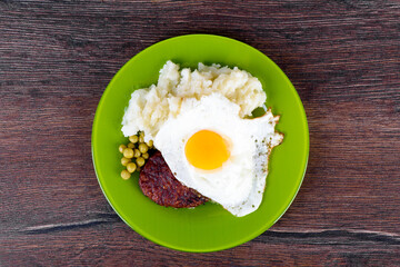 Top view of appetizer of cutlet with fried egg, mashed potatoes and green peas, on a plate.