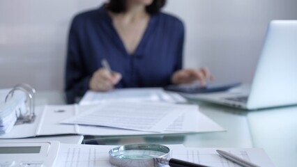 Magnifying glass, white calculator, pen and documents are lying on accountant's desk. Businesswoman wearing blue dress is working with documents on the background. Business and audit concept