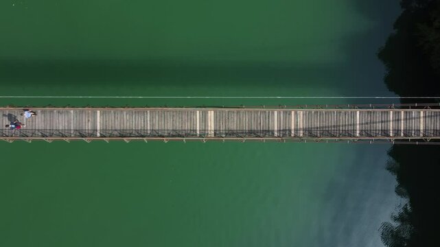 Top down view of two people walking on a narrow pedestrian bridge over a lake with green water. Aerial view natural environment at Arsi&egrave; lake, Italy
