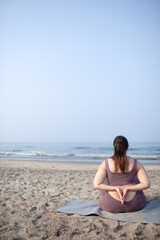 plus size girl does yoga on  the sea beach to lose weight and keep fit. The woman is wearing sports clothes, a purple top and leggins.