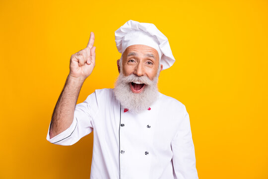 Cheerful senior chef in professional uniform pointing up with excitement on a vibrant yellow background, showcasing culinary skills