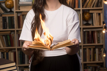 A woman in a white shirt holds a burning book in a cozy library, surrounded by shelves and warm...
