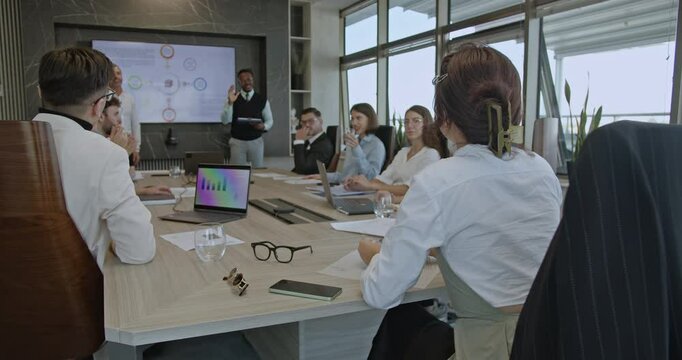 Multicultural colleagues discuss ideas in a business meeting, led by a speaker presenting on a large screen. Teamwork and collaboration in a diverse office setting.