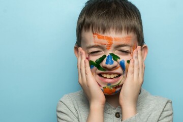 A joyful boy with colorful paint on his face holds his cheeks, laughing against a light blue...